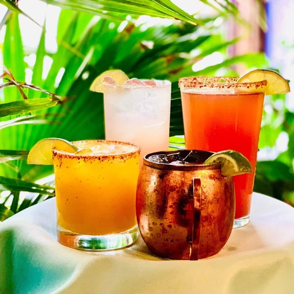Four colorful cocktails, including a Moscow Mule, on a white table with green foliage in the background.